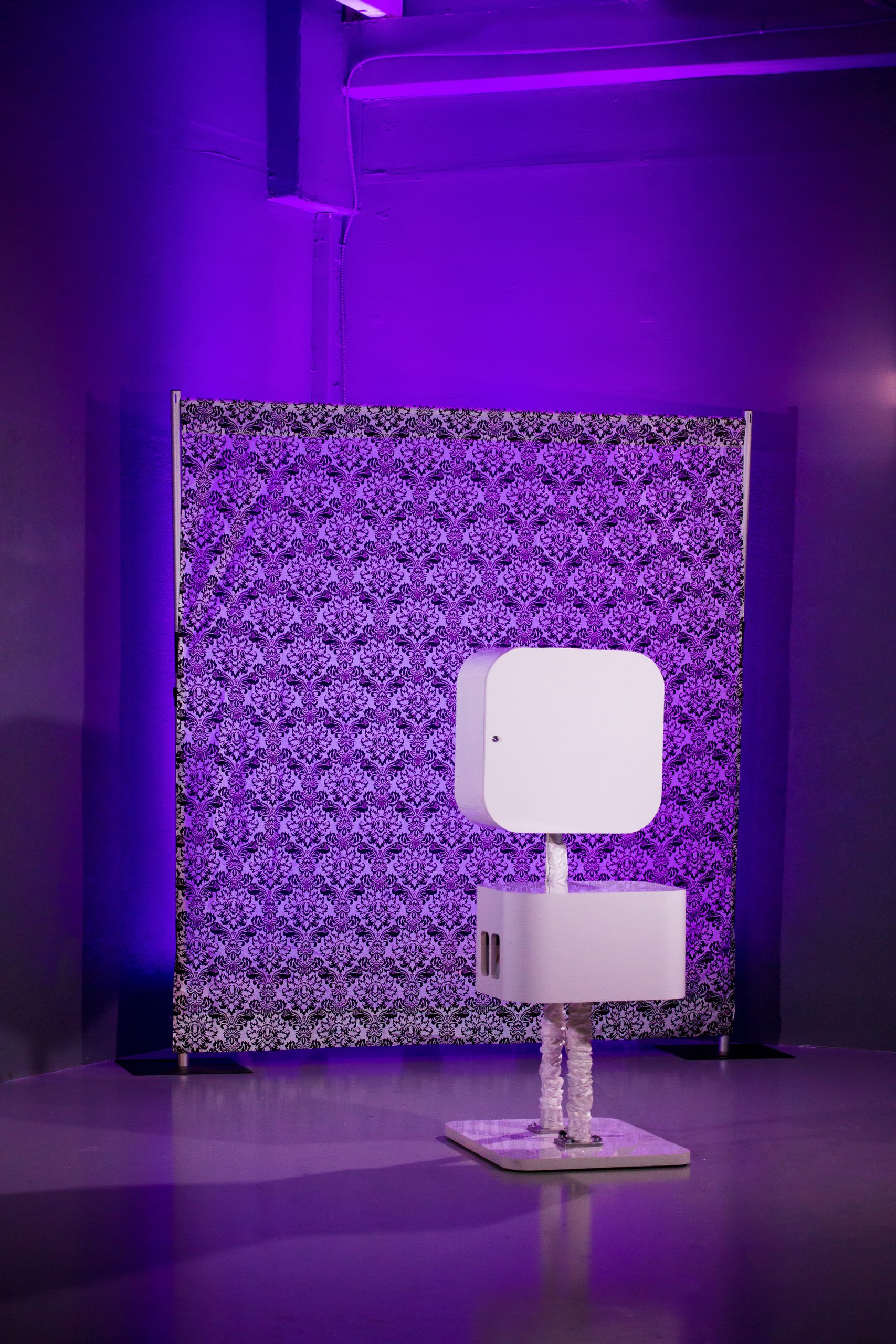 Miami Photo Booths A photo booth with a modern white design stands in front of a patterned backdrop. The setting is illuminated by purple lighting, creating an atmospheric ambiance.