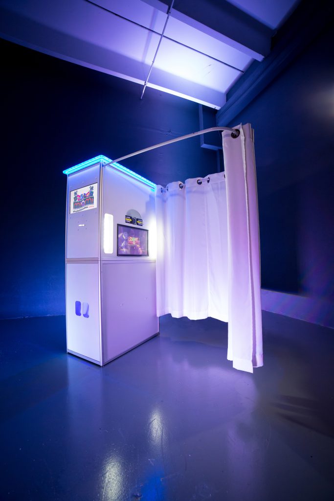 Miami Photo Booths A photo booth with bright blue LED lighting is set against a dark blue wall. It features white curtains partially open, revealing the interior. The booth stands on a smooth, reflective floor, giving a modern, sleek appearance.